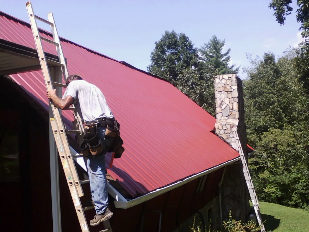 Roofer on a ladder beside a red metal roof and stone chimney — Soffit & Fascia Repair by Roofing Brothers in Ukiah